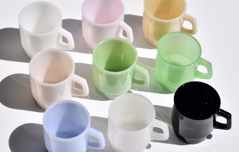 A classic white milk glass mug sitting on a wooden table next to a pot of coffee.
