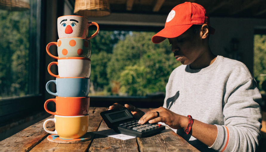 A person using a calculator next to a stack of custom-designed mugs, planning their business profits